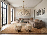 A modern living room featuring a taupe sectional sofa, two shaggy faux-fur ottomans, a glass coffee table, and a large textured area rug. The room has white wainscoting, a geometric wall art piece, and black-framed windows with beige curtains.